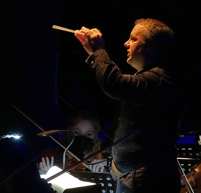 Profile view of conductor with baton conducting orchestra in front of black background