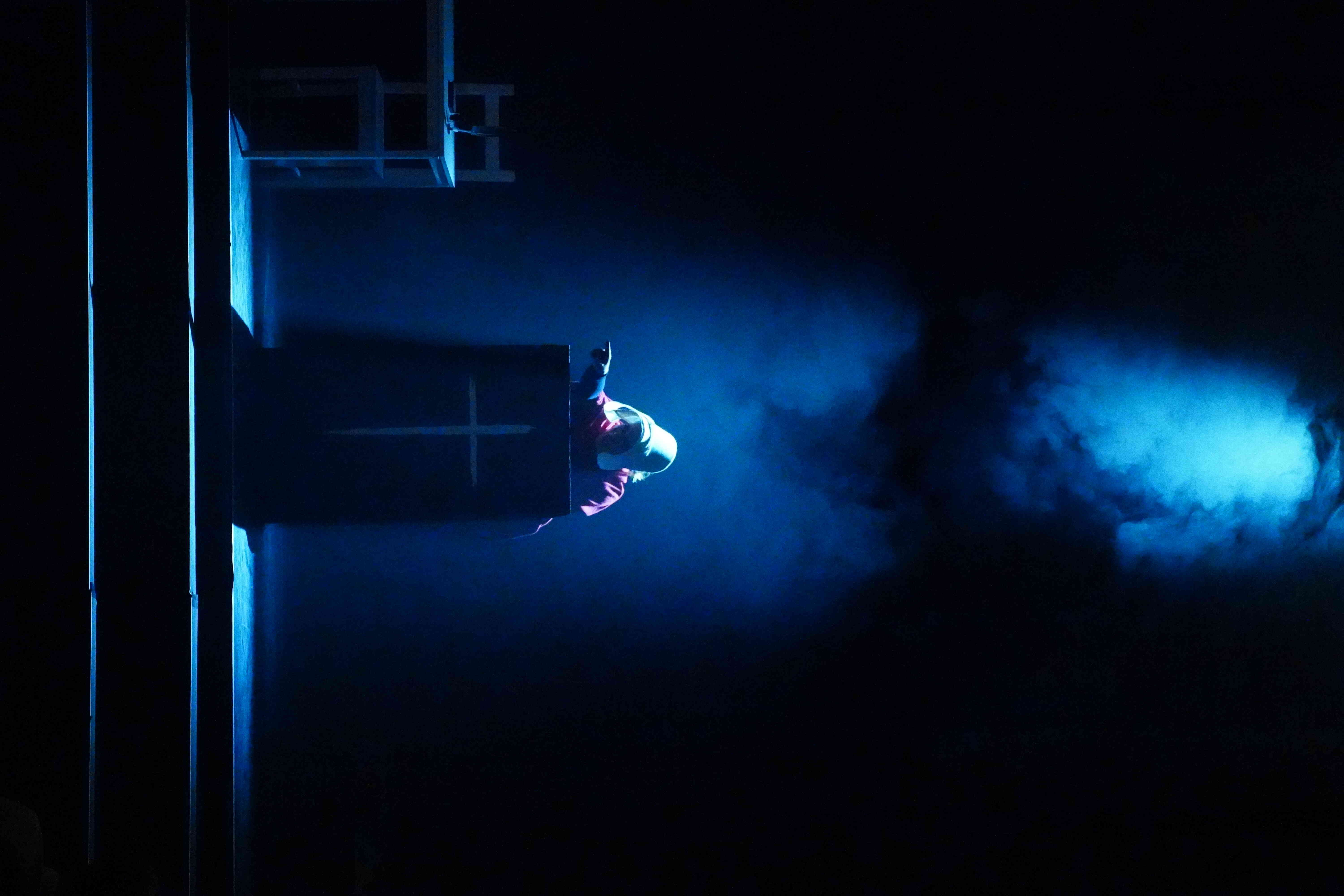 Portrait image with blue light illuminating white fog in the darkness of the stage. Single woman pointing from a podium with a cross on it.